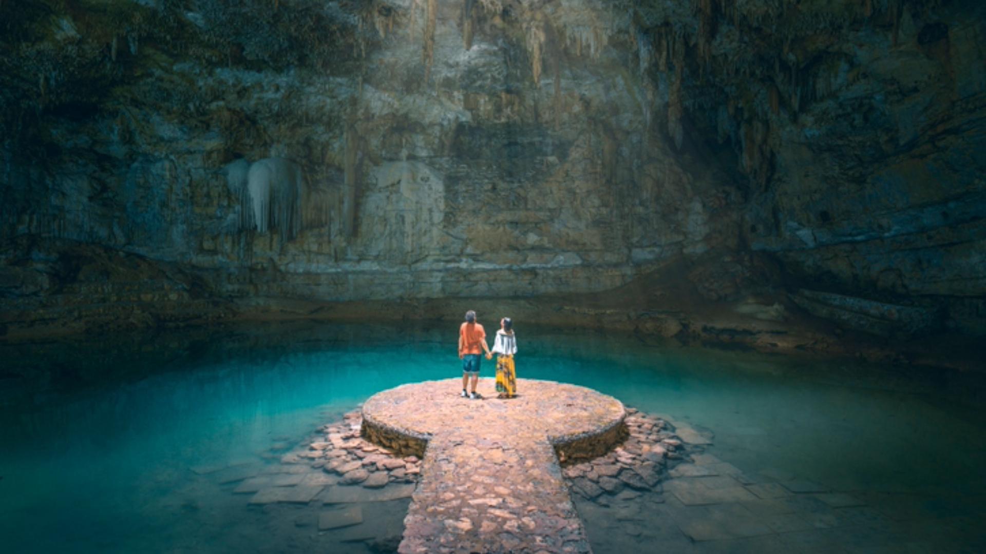 A couple stands on a circular stone platform surrounded by the turquoise water of a cenote or subterranean cave, with stalactites hanging from the cavern ceiling.