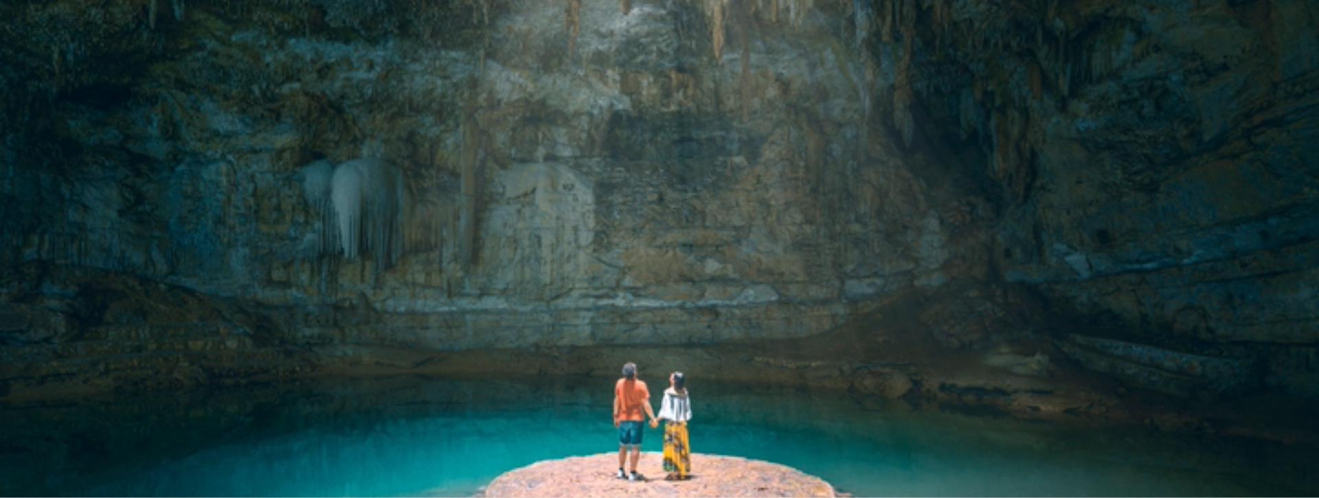 A couple stands on a circular stone platform surrounded by the turquoise water of a cenote or subterranean cave, with stalactites hanging from the cavern ceiling.