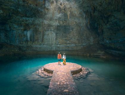 A couple stands on a circular stone platform surrounded by the turquoise water of a cenote or subterranean cave, with stalactites hanging from the cavern ceiling.