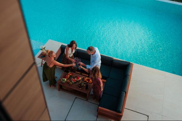 An overhead view of four adults clinking wine glasses around a coffee table loaded with snacks, sitting on a modern outdoor sofa beside a large, bright blue swimming pool at a resort.