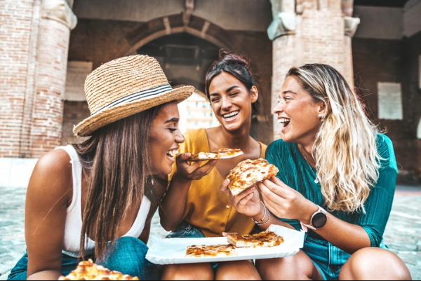 Three young women laughing and sharing pizza outdoors in an Italian piazza, representing a travel advisor-planned culinary trip or travel gift.