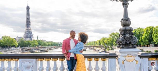 A romantic image of a couple embracing on a Parisian bridge with the Eiffel Tower and the Seine River in the background, promoting Black Friday travel gifts for couples.