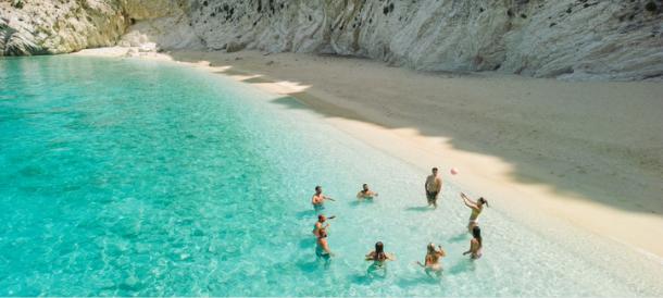 A group of eight friends or family members playing with a ball in the shallow, clear turquoise water of a secluded cove. Ideal image for a Black Friday group travel offer.