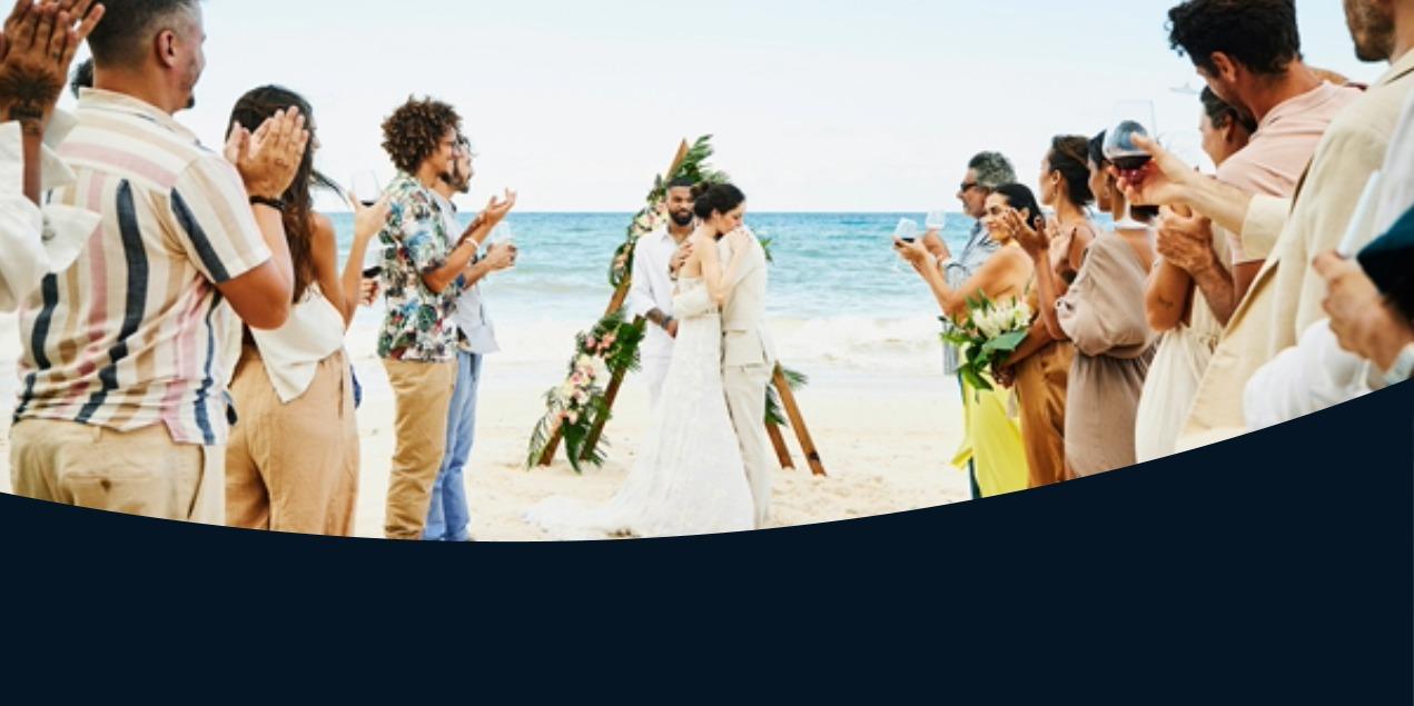 A couple getting married on a beach is surrounded by wedding guests who are clapping and toasting them.
