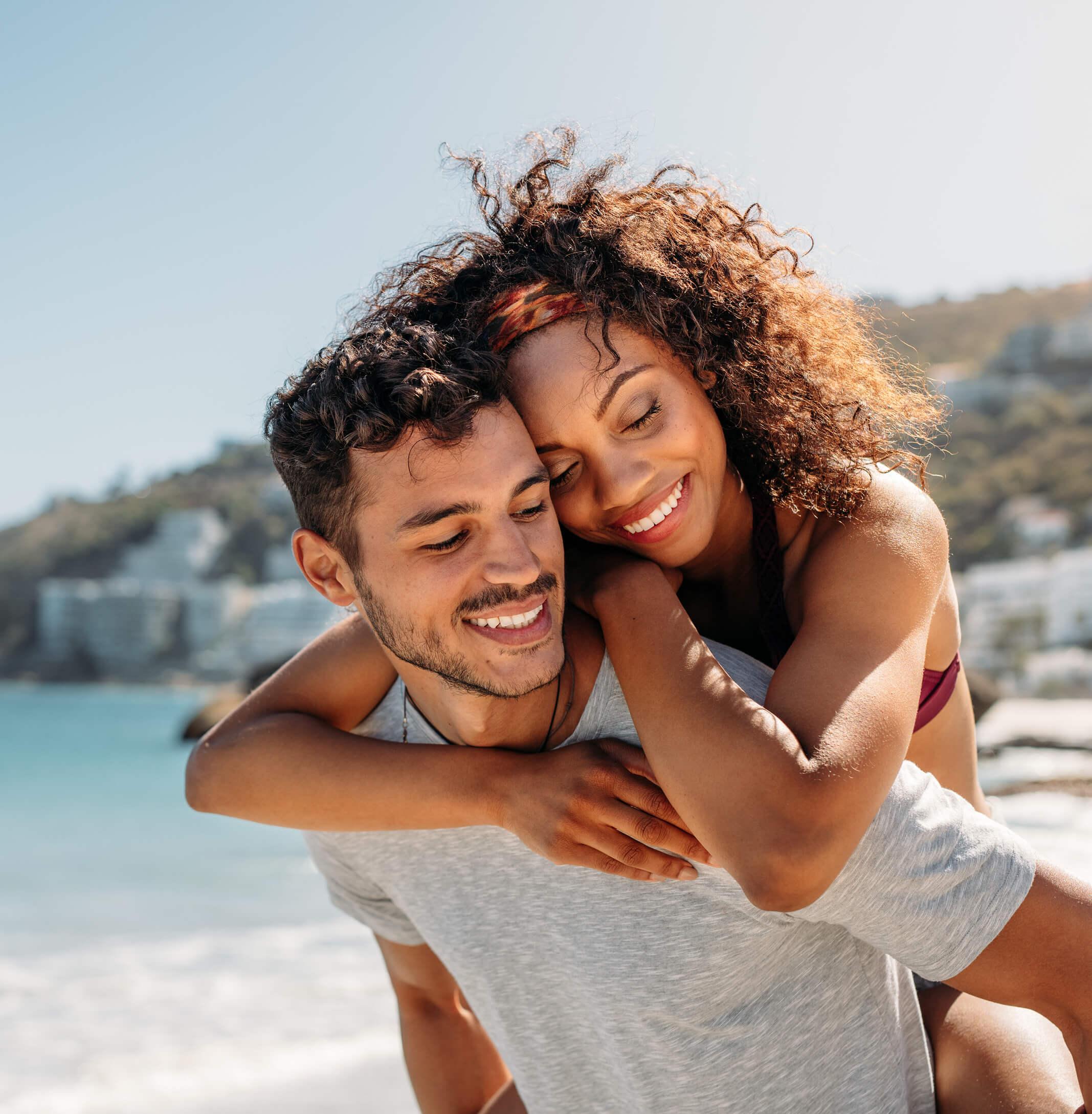 A woman hugs her partner from behind on a beach