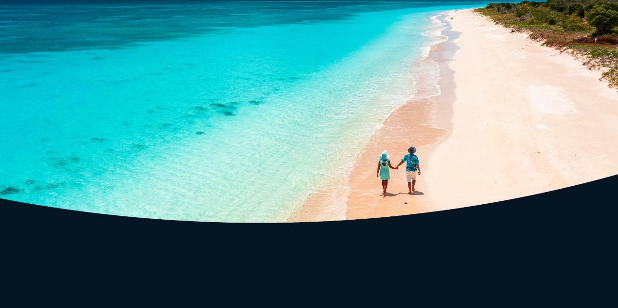 A romantic image of two people walking down a wide, pristine tropical beach where the clear turquoise water meets the shore.